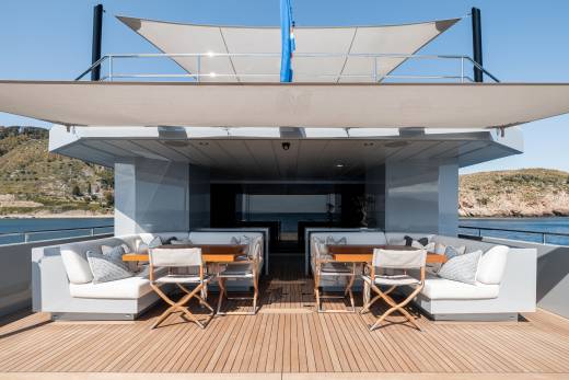 A modern, sleek yacht with a spacious outdoor deck featuring comfortable seating arrangements and a shaded canopy, set against a backdrop of a scenic coastal landscape.