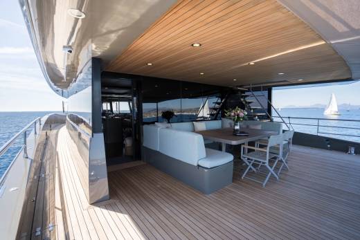 A luxurious yacht deck with a seating area and dining table in the foreground overlooks a calm sea with sailboats in the background.