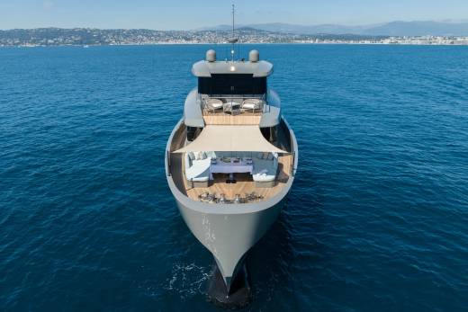 A sleek, modern yacht with an outdoor dining area in the foreground sails on calm blue water with a distant coastline in the background.