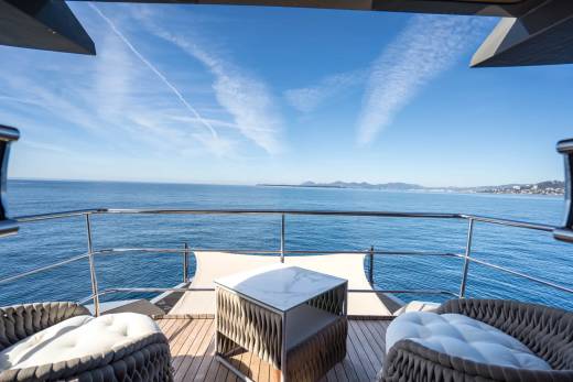 Two plush chairs and a modern table sit on a yacht's deck, overlooking a vast blue ocean under a clear sky with wispy clouds.