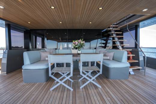 A spacious yacht deck with a dining table and seating in the foreground, and a staircase leading to the upper deck with a view of the ocean in the background.