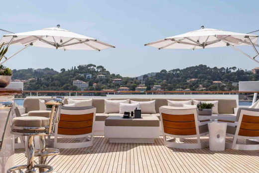 Two large white umbrellas shade a luxurious outdoor seating area on a yacht's deck, with a lush, green hillside dotted with houses in the background.