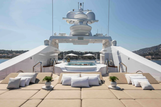 A luxurious yacht with plush seating in the foreground is anchored in a calm bay with a lush, green hillside in the background.