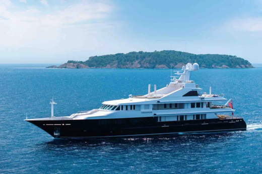 A sleek, dark-hulled yacht cruises on calm blue water with a lush, green island in the background under a hazy sky.
