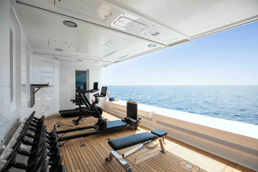 A well-equipped gym with weights, a treadmill, and a rowing machine sits on a wooden deck overlooking the calm blue ocean under a clear sky.