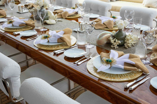 A long, polished wooden table is set for a formal meal with white chairs and elegant place settings in the foreground, while a blurred background suggests an outdoor setting with plush seating.