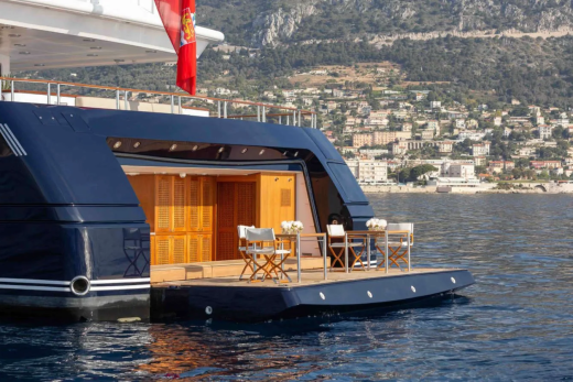 A luxurious yacht with an open deck featuring a dining set is anchored in calm blue water with a hillside town and mountains in the background.