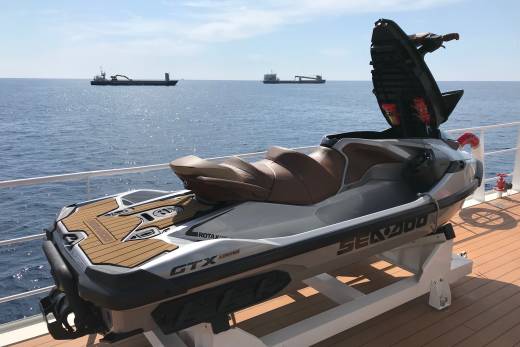 A silver and brown jet ski rests on a deck in the foreground, with two large ships sailing on a calm blue sea under a bright sky in the background.