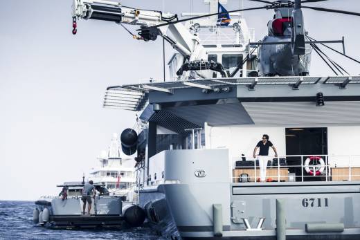 A man in white pants stands on the deck of a large yacht in the foreground, while a helicopter is visible on the upper deck of another vessel in the background.