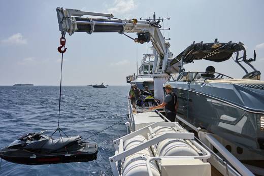 A jet ski is being lowered by a crane from a boat in the foreground, with a luxury yacht and a hazy horizon in the background.