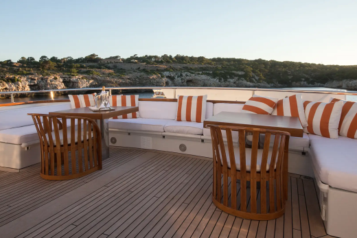 Wooden chairs and white cushioned seating with orange striped pillows are arranged around tables on a yacht's deck, with a rocky, tree-covered coastline visible in the background under a clear sky.