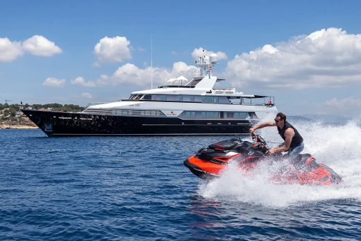 A man rides a jet ski through choppy water in the foreground, with a large yacht anchored in the background near a coastal town.