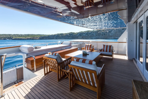 A luxurious yacht deck with comfortable seating and a table in the foreground overlooks a serene bay with a tree-covered coastline in the background.
