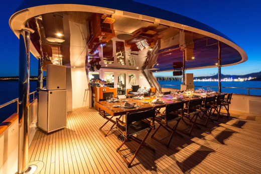 A long dining table set for a meal on a yacht's deck is illuminated against a twilight sky with a distant coastline.