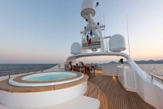 A circular jacuzzi sits on the wooden deck of a luxurious yacht, with the ocean and distant hills visible in the background under a soft twilight sky.