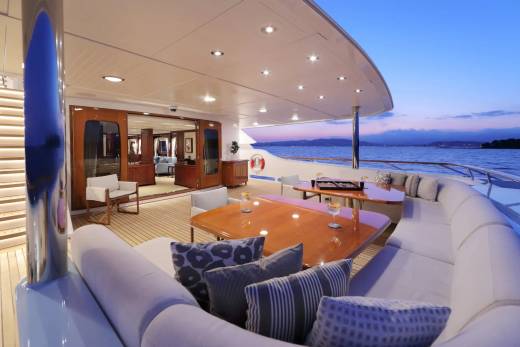 A luxurious yacht deck with plush seating and dining tables in the foreground overlooks a calm sea at dusk.