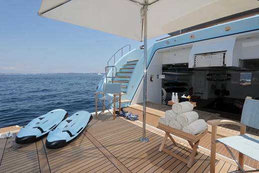 Two blue sea scooters rest on the wooden deck of a yacht in the foreground, while the background reveals a staircase leading up to the yacht's upper deck and the calm blue sea.