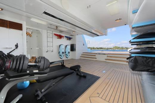 Dumbbells and a weight bench are in the foreground of an outdoor gym on a yacht, with a scenic view of water and trees in the background.