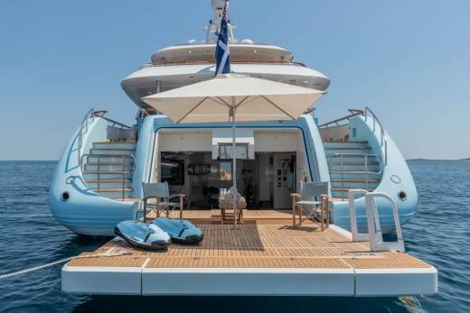 Two jet skis rest on the wooden deck of a light blue yacht, with a large white umbrella providing shade in the foreground, while the calm blue sea stretches to the horizon in the background.
