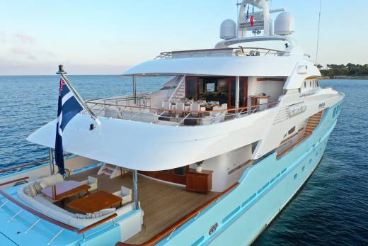 A large, light blue yacht with a wooden deck and outdoor seating in the foreground is anchored in calm blue water with a hint of land in the background.