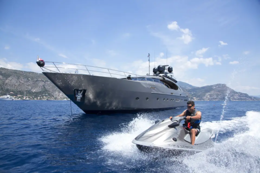 A man on a jet ski speeds across the water in the foreground, with a large, dark yacht anchored behind him and a lush, green coastline in the background.