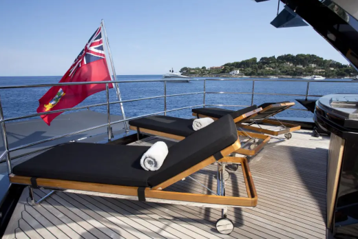 Two black-cushioned lounge chairs with rolled towels sit on a yacht's deck in the foreground, while a red flag flies above a calm blue sea with a distant coastline in the background.