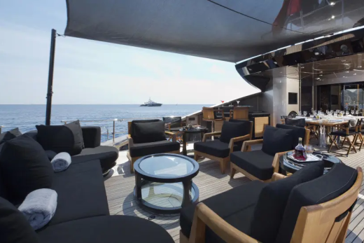Plush black outdoor seating and a dining table are on a yacht's deck, with a calm sea and distant ship in the background.