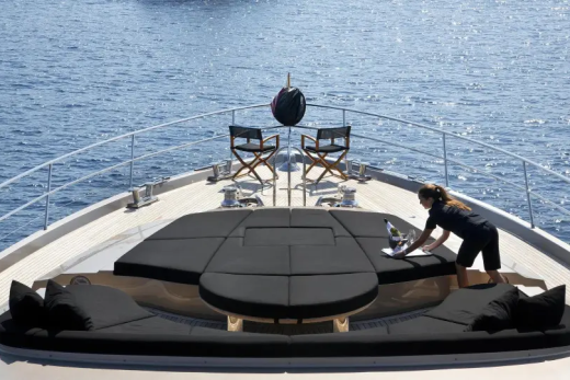A person cleans a plush, dark seating area on the bow of a yacht, with two director's chairs and the calm blue sea in the background.