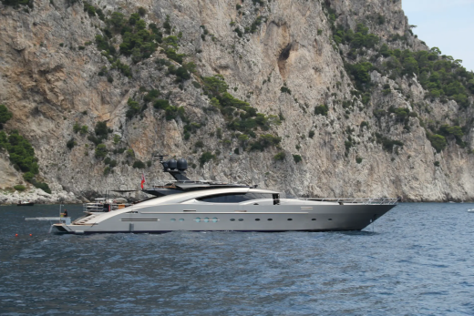 A sleek, modern yacht floats on calm blue water in the foreground, with a towering, rugged cliff face rising steeply behind it.