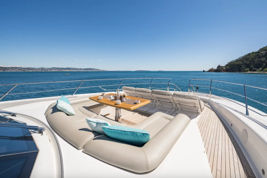A luxurious yacht's curved seating area with a table is in the foreground, while a calm blue ocean and a tree-covered coastline stretch into the background under a clear sky.