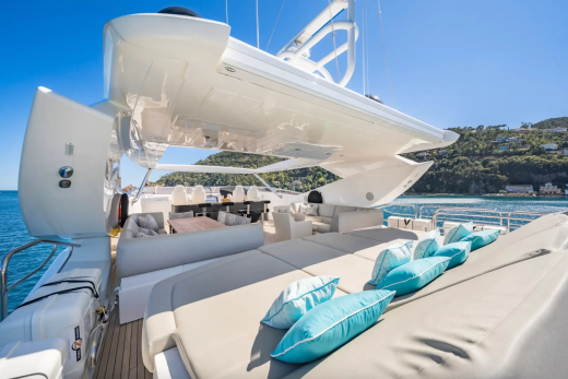 Plush turquoise cushions rest on a sun-drenched deck of a luxurious yacht, with a tranquil bay and lush green hills visible in the background.