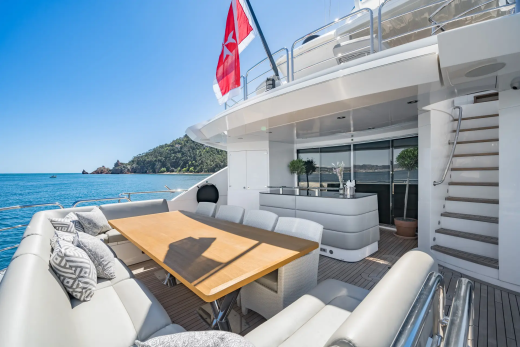 A luxurious yacht deck with a dining table and seating in the foreground, and a scenic coastline with a red flag in the background.