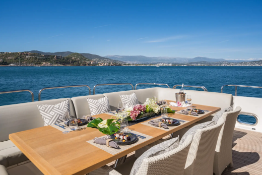 A beautifully set dining table on a yacht's deck overlooks a calm blue sea and a distant, sun-drenched coastline.