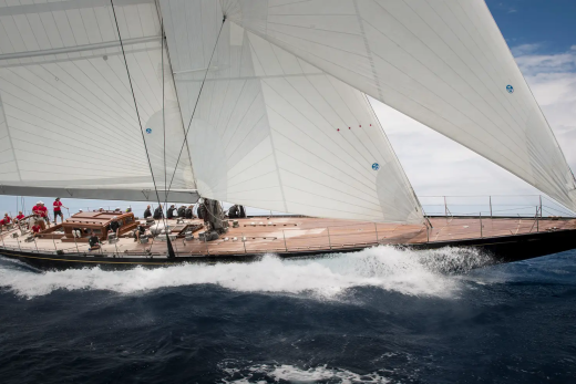 A majestic J-class yacht with full sails cuts through choppy blue water under a cloudy sky.