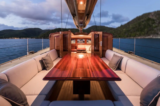 A polished wooden table and plush seating on a yacht's deck are set against a backdrop of calm water and tree-covered hills under a twilight sky.