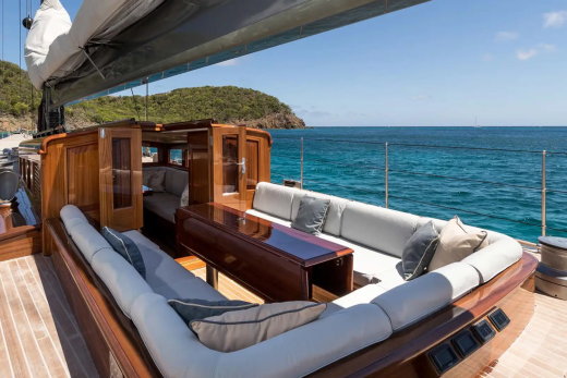 A luxurious yacht with plush seating in the foreground is anchored in a clear blue ocean with a lush green island in the background.