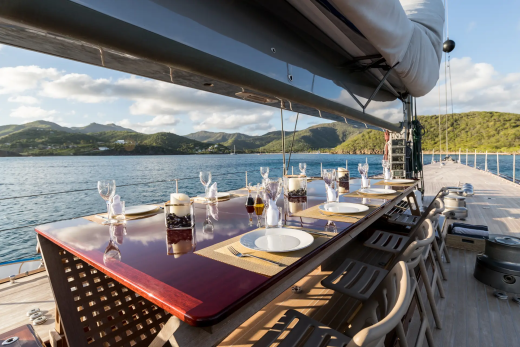A long table set for dinner on a yacht's deck overlooks a calm sea and lush green hills under a bright sky.