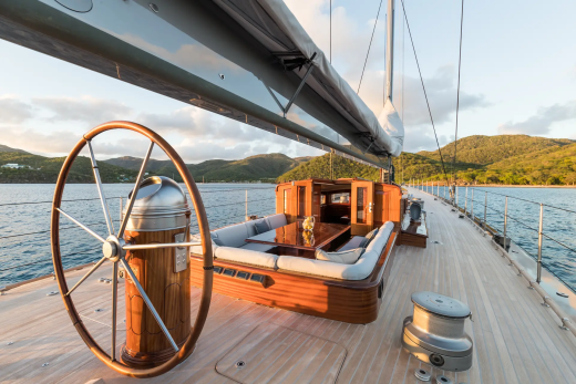 A wooden ship's wheel and seating area are in the foreground, with a lush, green coastline and calm blue water in the background.