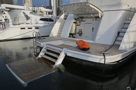 A white yacht with a lowered swim platform and an orange rescue device in the foreground is docked next to another white yacht with city buildings in the background.