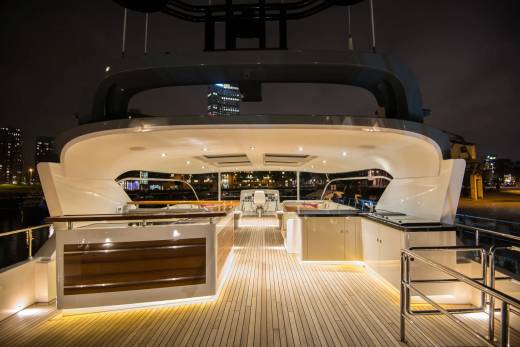 A luxurious yacht deck with illuminated wooden flooring in the foreground is set against a backdrop of a city skyline at night.