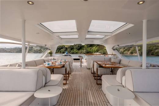 Luxurious white sofas and dining tables fill the foreground of a yacht's deck, while the background reveals a tranquil bay with lush green hills.