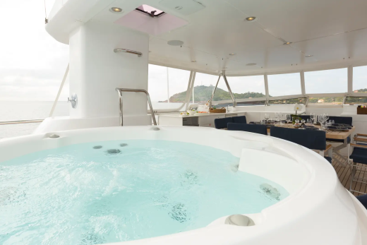 A bubbling hot tub in the foreground overlooks a dining area and a scenic coastal view in the background.