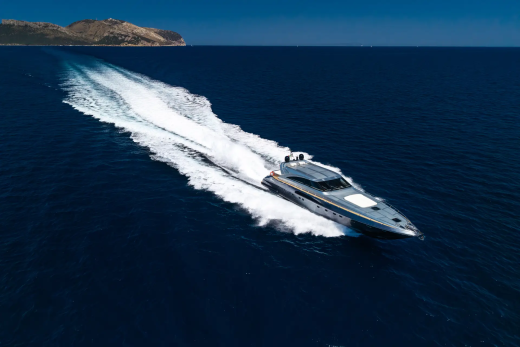 A sleek, modern yacht speeds across the deep blue ocean, leaving a white wake, with a distant, sun-drenched coastline under a clear sky.