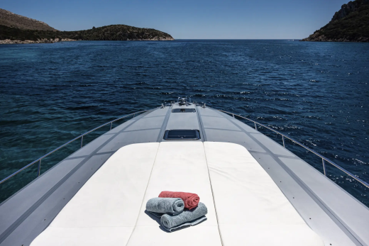 A sleek yacht with folded towels on its deck glides through calm blue water towards a distant, hilly coastline under a clear sky.