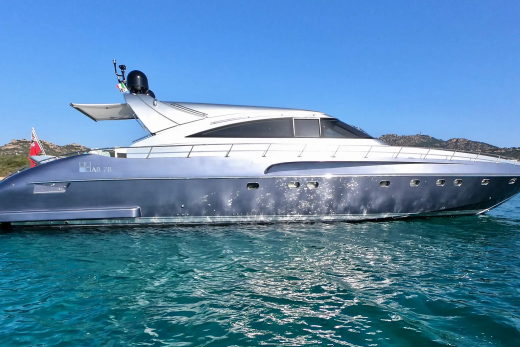 A sleek, silver yacht floats on calm, turquoise water in the foreground, with a lush, green coastline under a clear blue sky in the background.