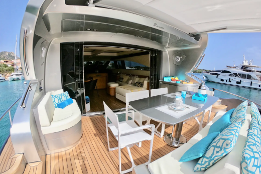 A luxurious yacht deck with a dining table and seating in the foreground, and other yachts docked in a clear blue bay in the background.