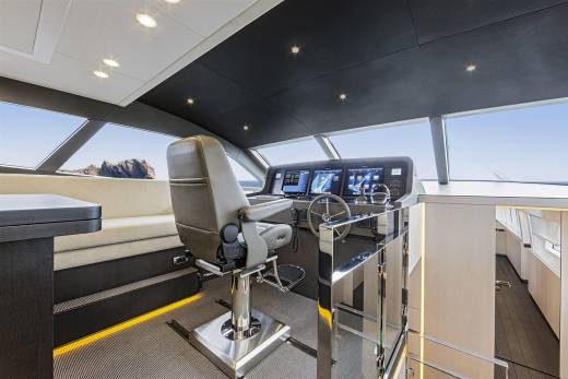 A luxurious yacht's helm station with a captain's chair and advanced navigation screens is in the foreground, while a scenic ocean view with rocky islands is visible through the large windows in the background.