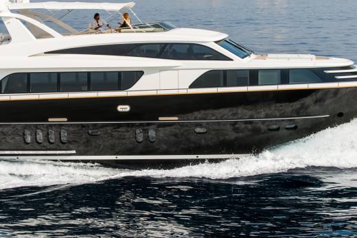 A sleek, dark-hulled yacht cruises through choppy blue water, with two people visible on its upper deck against a hazy sky.