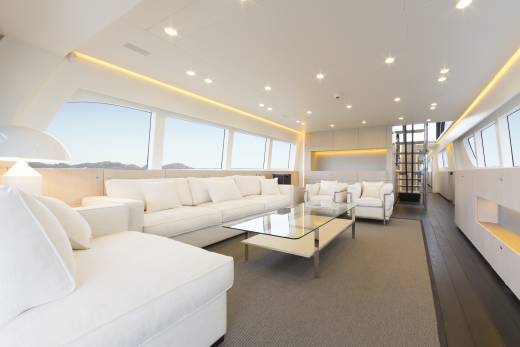 A spacious, modern white living area with plush sofas and a glass coffee table in the foreground is set against a backdrop of large windows offering views of a bright, outdoor landscape.