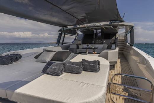 A spacious sun deck with plush cushions and towels is in the foreground, while a sleek, modern yacht with comfortable seating and a dark canopy is in the background, set against a backdrop of the sea and sky.